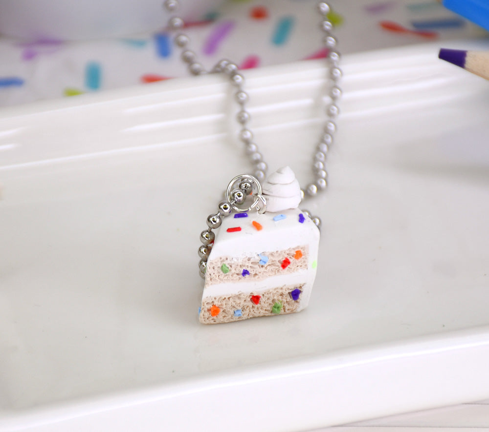 A miniature slice of white layered cake charm with sprinkles on a silver ball-chain necklace.