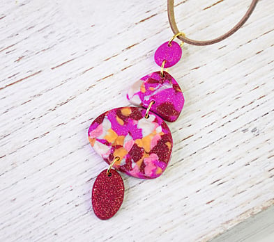 A handcrafted clay pendant necklace featuring multiple tiered organically shaped pieces in vibrant magenta , orange , and white with glitter accents.