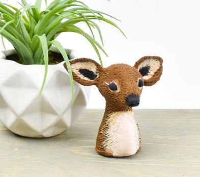 A handcrafted brown clay deer head figurine with a white chest standing on a light wood surface next to a white geometric planter.