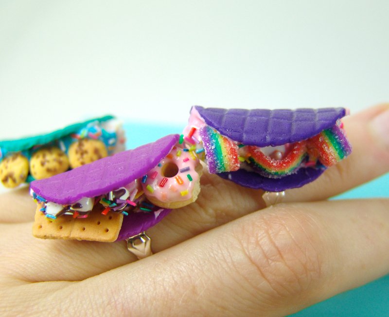 Three whimsical clay rings shaped like purple waffles or taco shells filled with miniature treats like a donut rainbow candy and a s'more.
